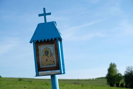A small roadside blue chapel with the Mother of God among green fields. Polish countryside,...