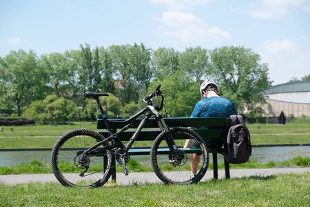 Cracow, Poland - circa May 2020: Young man sitting backwards on a bench by the Vistula river on a sunny summer day. There is a bicycle next to it.の写真素材
