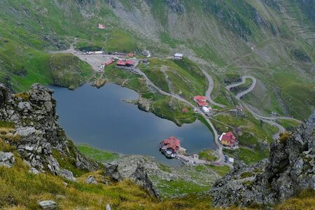 Romania, Romanian Carpathians - Fagaras Mountains, view from the ridge on the lake Balea Lac, houses around and the zigzag road (bends) of the Transfagarasan Highway roadの写真素材