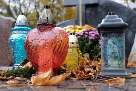 Colorful glass candles among dry autumn leaves on grave in cemetery on November day