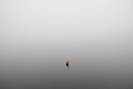 Single red buoy on surface of very calm lake in whiteness on foggy dayの写真素材