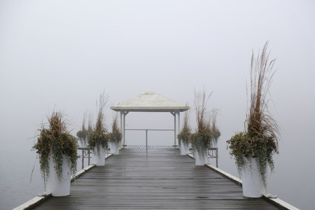 Romantic moody scenery with wooden pier with white gazebo by lake on calm misty dayの写真素材