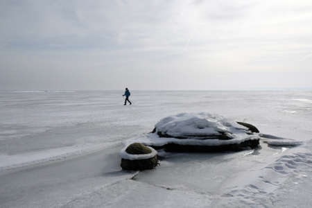 Ice-covered stones on the shore of a frozen sea during the thaw. Silhouette of walking person on frozen sea.の写真素材