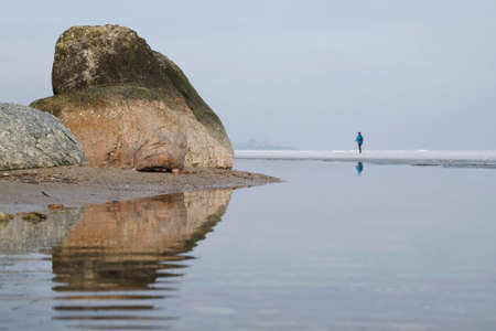 Silhouette of walking woman with poles on horizon of sea with very calm water. Interesting perspective - stones on shore are looking very big. Oslonino, Baltic Sea, Polandの写真素材