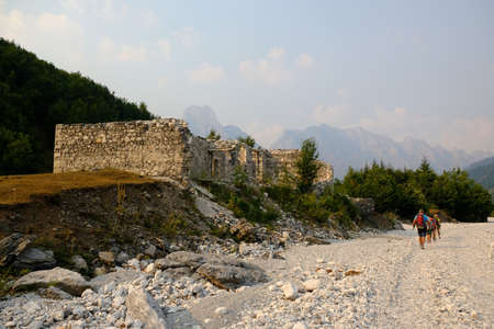 Group of tourists are walking down Valbona Valley in Albanian Alps, Albania. There is ruined building next to road.のeditorial素材