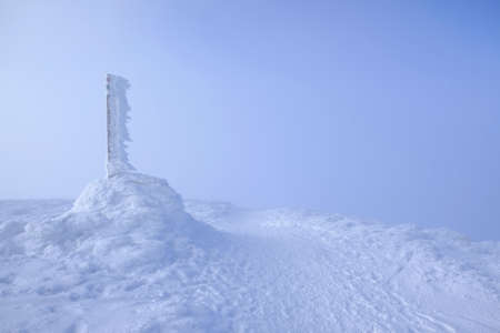 A frozen pole marking a trail in the mountains by a path on a foggy winter day. Babia Gora massif,...