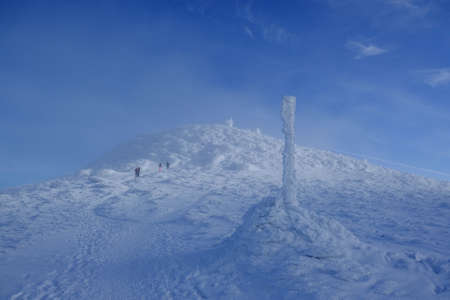 Frozen pole marking trail in mountains by path on misty and sunny winter day in Babia Gora massif. Silhouettes of people climbing to Babia Gora peak in background. Diablak, Beskid Zywiecki, Polandの写真素材