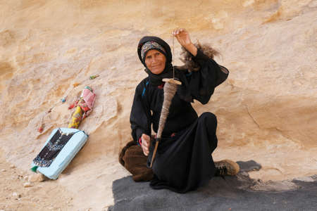 Little Petra (Siq al-Barid), Jordan - circa May 2022: Old Jordanian woman in traditional dress is making camel wool threads on the spindle in Little Petraのeditorial素材