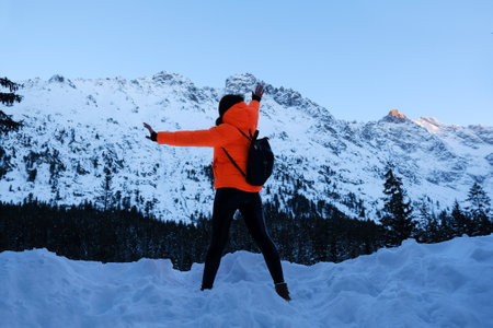 Woman in winter jacket in intense orange color is standing with raised arms on trail to Morskie Oko (Sea Eye) in Tatras. Tatra National Park, Polandの写真素材