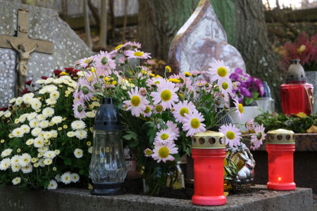 Grave with glass candles and flowers in pots among dry autumn leaves in cemetery on November dayの写真素材