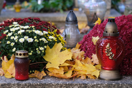 Grave with glass candles and flowers in pots among dry autumn leaves in cemetery on November dayの写真素材