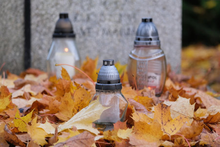 Grave with glass candles among dry autumn leaves in cemetery on November dayの写真素材