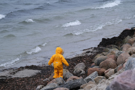 Little child in a yellow waterproof warm suit is standing on the beach by the sea. Baltic Sea, Polandの写真素材