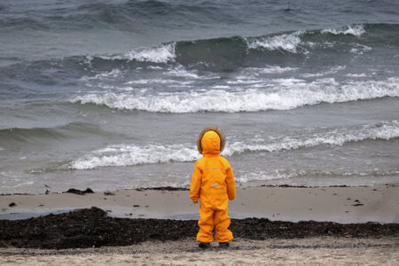 Little child in a yellow waterproof warm suit is standing on the beach by the sea. Baltic Sea, Polandの写真素材