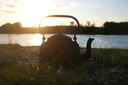 Title: An old metal kettle stands on the grass by the lake at sunset.の写真素材