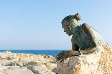 Paphos, Cyprus. A sculpture of a woman paying tribute to Aphrodite. Rocks on the seaside boulevard.の写真素材