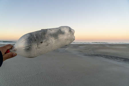 Hand holding plastic bottle by the ocean at sunset