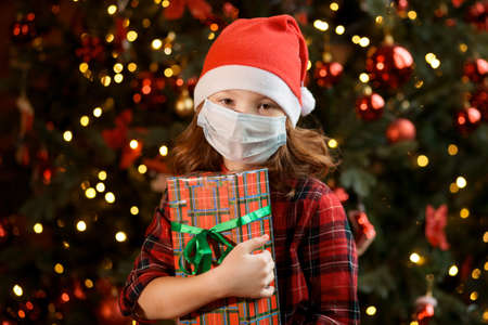 Girl in Santa hat and medical mask near Christmas tree with giftの写真素材