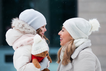 Little daughter looking at mother and holding teddy bear in winter cityの写真素材