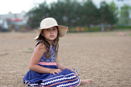 Little sad girl sitting in summer hat and beautiful dress on the beach