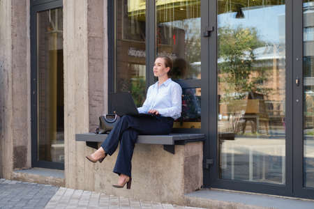 Portrait of business young woman, smiling while sitting on bench outdoors with laptopの写真素材
