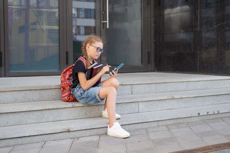 redhead girl in eyeglasses with books and backpack typing on phoneの写真素材