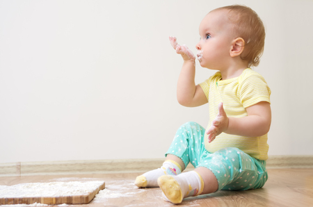 A little cute baby-girl with on her head is sitting on the kitchen floor, eats flour, she is heavily soiled with wheat flourの写真素材