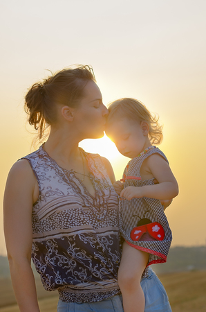 Happy family: young beautiful woman kisses on the forehead of her little cute daughter, stand in a wheat orange field on a sunny summer day. The attitude of parents and children. Nature in the country.の写真素材