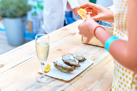 A woman eats fresh oysters. a wooden table, a plate with three oysters, salt, a woman's hands, a glass of champagne, a phone, an outdoor food market.の写真素材