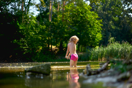 Adorable curly toddler girl wearing in pink shorts, playing at a river shore splashing water on sunny summer dayの写真素材