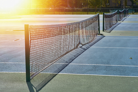 Sunshine over a tennis court in a public park. Tennis courts with blue surface and mesh fencesの写真素材