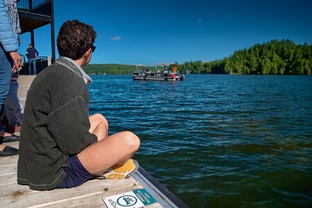 Lake Massawippi, Quebec, Canada -- September 8, 2018: young guy awaits sitting on the pier ferry to Ayers Cliff, Lake Massawippi, the eastern city of Magog, Quebec, late summer, Travel on Canada.のeditorial素材