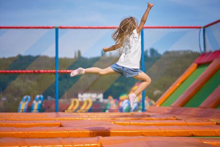 Little pretty girl having fun outdoor. Jumping on trampoline in children zone. Happy girl jumping on the yellow trampoline in Amusement park. girl on a trampoline jumps like a ballerinaの写真素材