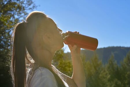 Attractive young blonde in a white T-shirt drinking water from a bottle, standing in the mountains...