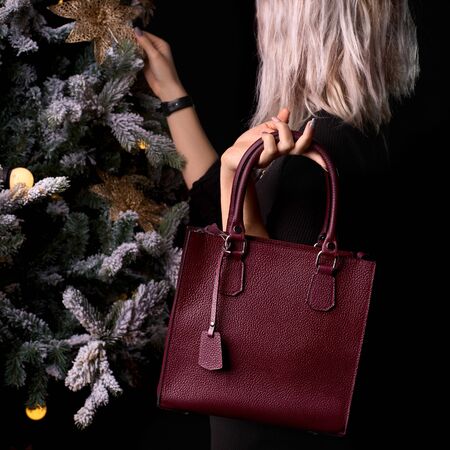 Modern burgundy leather bagin a female hand. girl near the christmas tree, 1:1 copy space for text and design. Accessory, studio shot.の写真素材