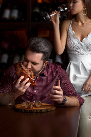 The guy eagerly eats a pork knuckle. Lady sitting on a couch, with a glass of red wine, on a dark background in a restaurant.の写真素材