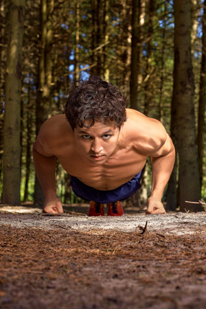 Sportsman with a naked torso and a focused face is ready to push up, in the forest against the...