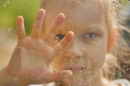 little girl looks out the wet window. Raindrops on the glass. Forest outside the window. Selective focus, Close up.の写真素材