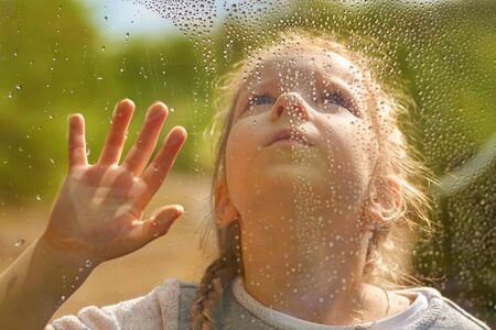 little girl looks out the wet window. Raindrops on the glass. Forest outside the window. Selective focus, Close up.の写真素材