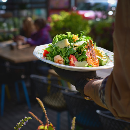 Fresh summer salad with shrimps in the hands of the waiter on the terrace, close up. Healthy food concept.の写真素材