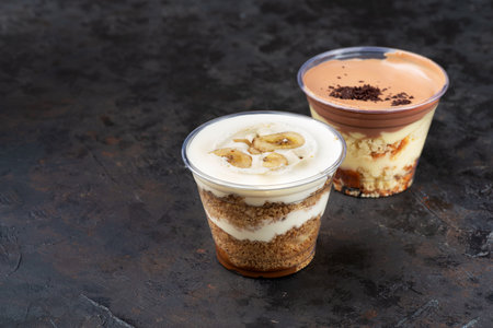 Fast food delivery. Banoffee pie dessert in cup with fresh banana on wooden background, selective focus, copy space, Takeaway boxの写真素材