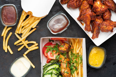 Fast food - box of crispy grilled chicken legs with Greek souvlaki and French fries on a dark stone...