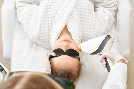 Young beautiful woman on beauty cosmetology procedure in spa salon. Therapist beautician makes a laser treatment to young womans face at beauty SPA clinic.の写真素材