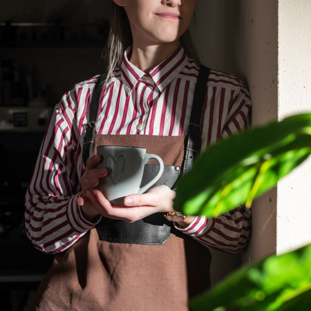 Young barista girl near a modern window enjoys a cup of coffee, looking outside, good morning, working day, free from work and duties. copy spaceの写真素材