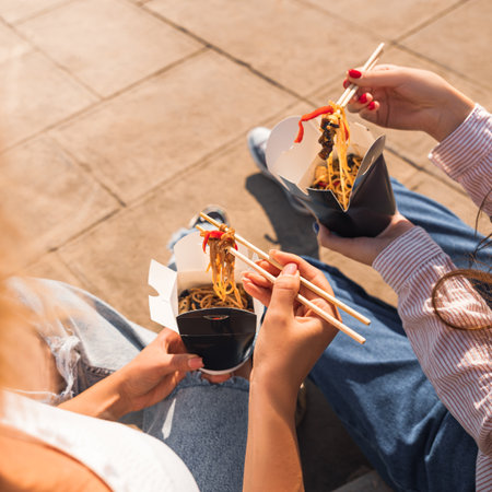 young girls, girlfriends eat asian food with wok noodles, using chopsticks, taking out takeaway food, paper box with takeaway noodlesの写真素材