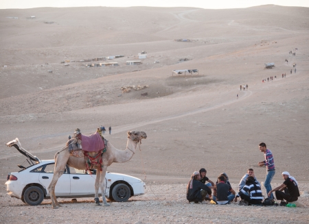 A group of Bedouins sitting beside their vehicles in the Judea desert  On the background a tourist group goes up a twisting road by scattered hovels のeditorial素材