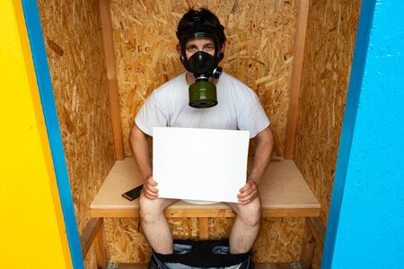 Funny man in a gas mask sitting in a stinking toilet. In the hands of a man a white banner with a copy space. The concept of bad smell the toilet. Wooden washroom countryside outhouseの写真素材