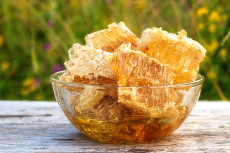 Honeycomb with golden honey in a glass bowl. Orange honey on an old rustic wooden table. The sun is shining on the honeycomb. Against the background of green grass, flowers bloom.の写真素材