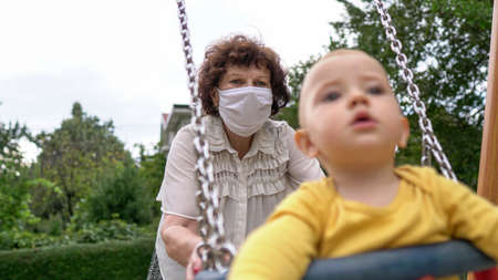 A woman with medical mask swinging a baby on a swing. Covid-19, coronavirus.