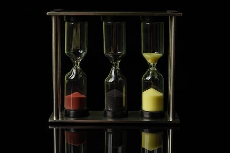 Three hourglasses with colorful sand stand on a black glossy glass, in which their reflection is visibleの写真素材